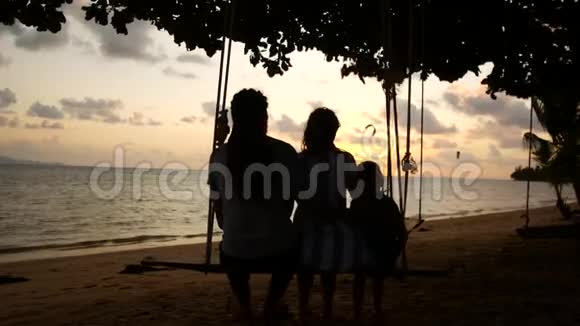度假的幸福家庭爸爸妈妈和宝宝骑在秋千上看海边的日落视频的预览图
