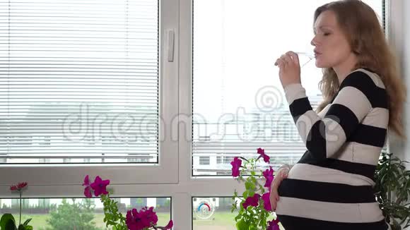 年轻怀孕模特的侧视图喝水透过百叶窗窗观看视频的预览图