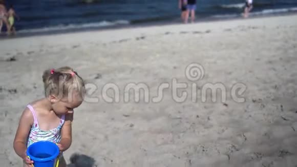 幼儿女孩拿着带水的玩具桶穿过沙滩模糊的人视频的预览图