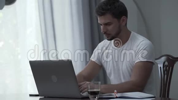 穿着白色t恤手提电脑和文件在家里或办公室喝咖啡的胡子男他累了视频的预览图