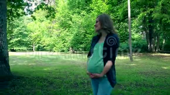 放松的孕妇在公园散步站在巨大的橡树树干附近视频的预览图