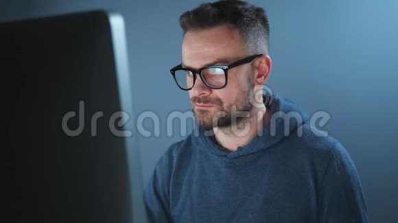 戴着帽衫和眼镜的长胡子男黑客在黑暗的办公室里的电脑上工作网络犯罪概念视频的预览图