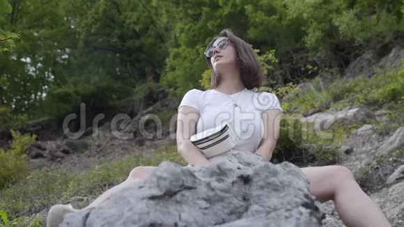 美丽的黑发女人独自坐在岩石和森林山背景旅游概念视频的预览图