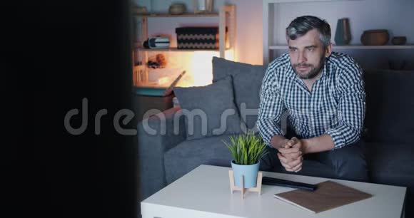 一个大胡子男人晚上坐在沙发上在黑暗的公寓里看电视视频的预览图