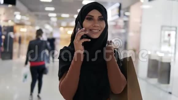 戴着专业化妆的年轻穆斯林女士穿着黑色头巾在现代购物中心的智能手机上聊天视频的预览图
