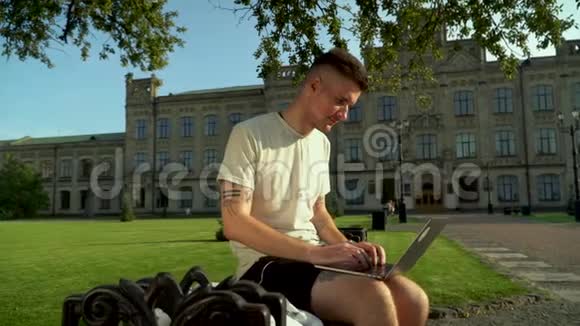 年轻忙碌的男人坐在漂亮的大楼前的长凳上拿着笔记本电脑工作学生在寻找视频的预览图