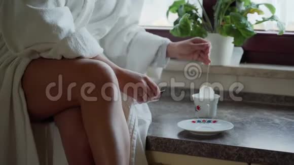 穿着白色浴袍的女人坐在舒适的厨房里的石桌上早上喝茶咖啡时间关闭视频的预览图