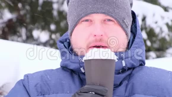 一个长胡子的人在冬天散步时喝咖啡从杯子里吹雪下雪视频的预览图