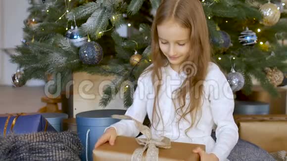 可爱的金发女孩收到了一个礼品盒包裹着工艺纸坐在圣诞树附近快乐的童年和圣诞节视频的预览图