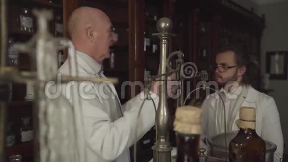老秃头药剂师拿着古老的烧瓶和试管和年轻的白人同事交谈视频的预览图