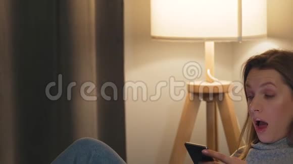 微笑的女人躺在舒适的房间里的沙发上用智能手机上网或和别人聊天视频的预览图