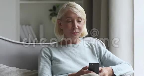 微笑的中年妇女喜欢使用智能手机坐在沙发上视频的预览图