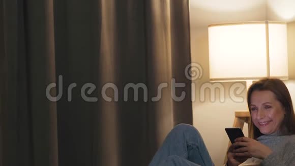 微笑的女人躺在舒适的房间里的沙发上晚上用智能手机上网放松和视频的预览图