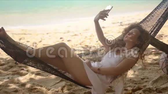 可爱的女人在沙滩上躺在吊床上用智能手机自拍视频的预览图