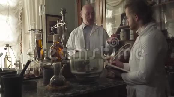 两名白人男子站在桌子旁手里拿着古老的烧瓶试管和化学设备年纪较大的药剂师视频的预览图