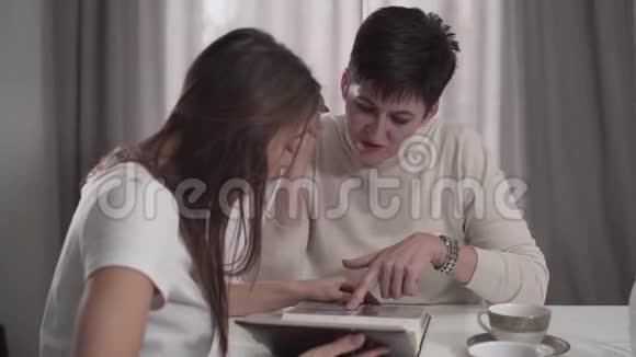 深色头发的年轻白种人女士与美丽的女儿在室内观看相册女人的故事视频的预览图