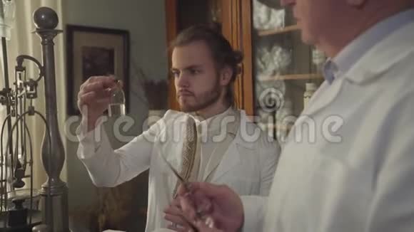 英俊的白人年轻人向年长的专业人士展示带有透明液体的瓶子两名药剂师的画像视频的预览图