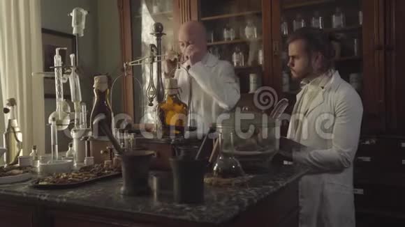 老白种人药剂师戴上眼镜向穿白色长袍的年轻人展示带有透明液体的瓶子新视频的预览图
