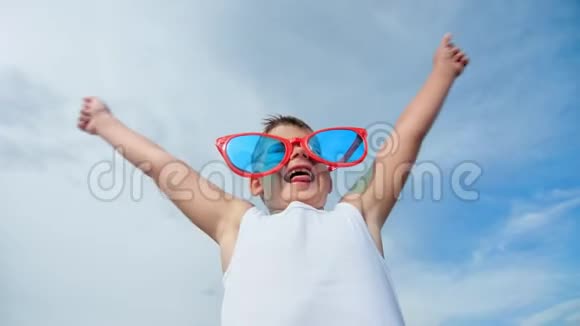 幸福的小男孩戴着大红色滑稽眼镜举起手在蓝天背景下有积极的情绪视频的预览图