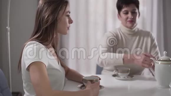 坐在桌边的年轻白人女孩的侧视图她漂亮的母亲正在往杯子里倒茶女儿视频的预览图