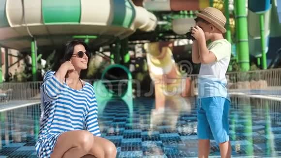 美丽的母亲在水上乐园水上滑梯背景下可爱的儿子用相机拍照视频的预览图