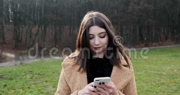 在森林附近的秋天公园年轻迷人的女人在绿色草坪上散步和看智能手机的肖像视频的预览图