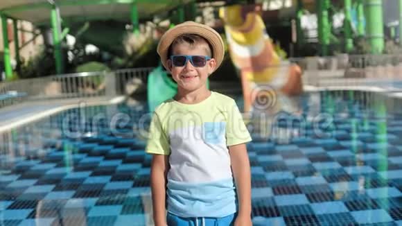 时髦的旅行者戴着太阳镜和帽子的婴儿指着游泳池附近的操场滑梯视频的预览图