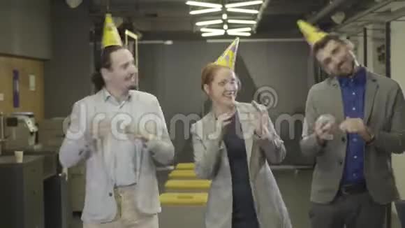 三个戴着派对帽的白种人办公室工作人员在合作空间走廊跳舞同事在公司里玩得开心视频的预览图