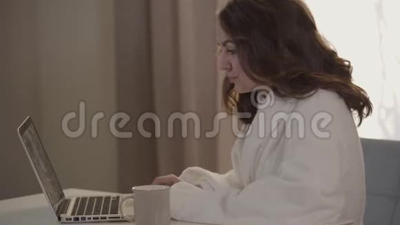早上上网的白种人女人的侧景特写穿着白色浴袍的年轻黑发女孩视频的预览图
