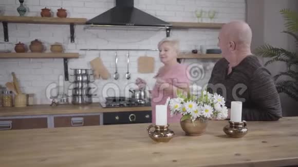 快乐的老白种人女士从厨房端来一杯白茶或咖啡和她可爱的丈夫交谈微笑视频的预览图