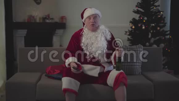 有趣的白人男性圣诞老人坐在沙发上看电视上的体育比赛资深快乐粉丝在家休息后视频的预览图