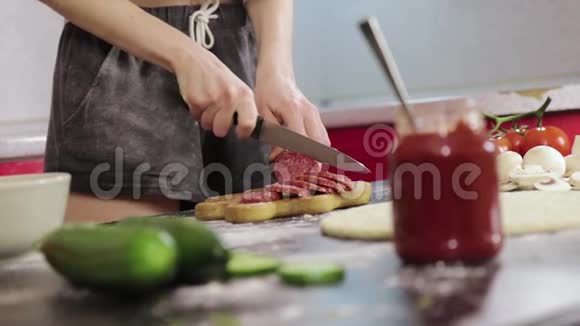 年轻女孩做披萨在木切割板上切烟熏香肠钝刀黑桌子视频的预览图