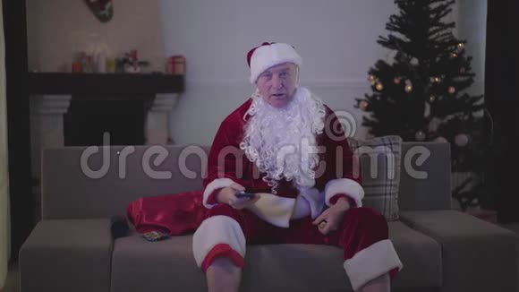 穿着圣诞老人服装的假白胡子老人在家里坐在沙发上看电视视频的预览图