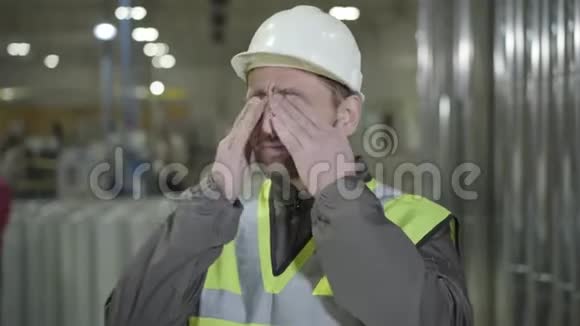 精疲力竭的白种人戴着头盔和背心在生产现场揉眼睛和伸展疲惫的年轻工人视频的预览图