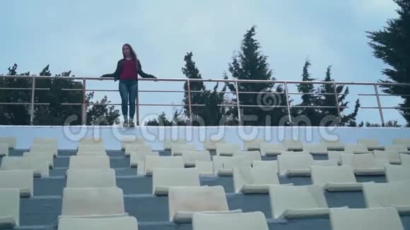 一个穿着自行车夹克和牛仔裤的美丽而热情的女孩独自站在一个空荡荡的体育场里靠在篱笆上视频的预览图