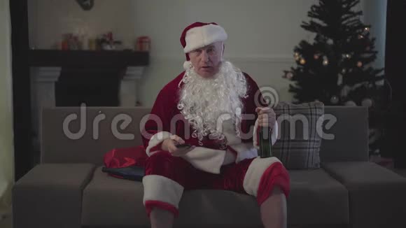 悲伤成熟的白种人穿着圣诞老人服装坐在沙发上喝啤酒看电视上等人休息视频的预览图