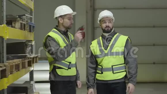 两个白人男子站在仓库里说话和打手势工人在生产现场计划时间表工厂视频的预览图