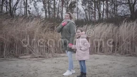 一边看着微笑的白种人女人和开朗的女孩站在秋天的海滩上看着远方等待的家人视频的预览图
