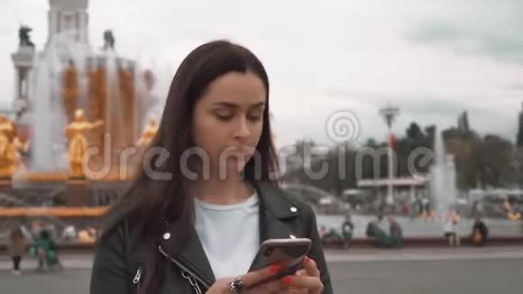 漂亮的女孩穿着黑色夹克和白色t恤在智能手机上输入短信关门视频的预览图