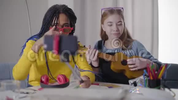 可爱的高加索女孩玩四弦琴她的非裔美国朋友录制自拍视频有趣的青少年玩得开心视频的预览图