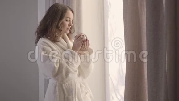 漂亮的白种人黑发女孩摇动咖啡杯喝一杯微笑穿着白色浴袍的年轻美女视频的预览图