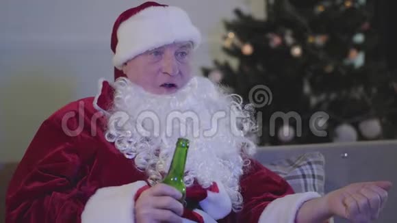 一个穿着圣诞老人服装咀嚼和喝啤酒的悲伤白种人的特写镜头坏圣诞老人坐在沙发旁边视频的预览图