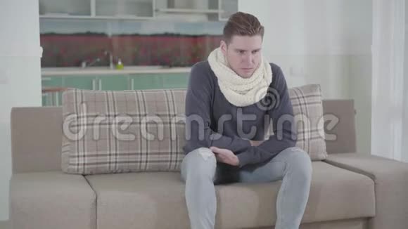 白人青年戴着白色围巾坐在沙发上咳嗽的肖像生病的家伙喉咙痛疾病视频的预览图