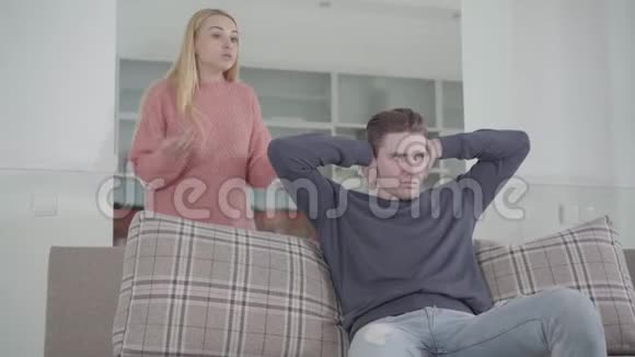 精疲力竭的白种人男人坐着双手紧闭着耳朵年轻漂亮的女人在骂他不满意的女孩视频的预览图