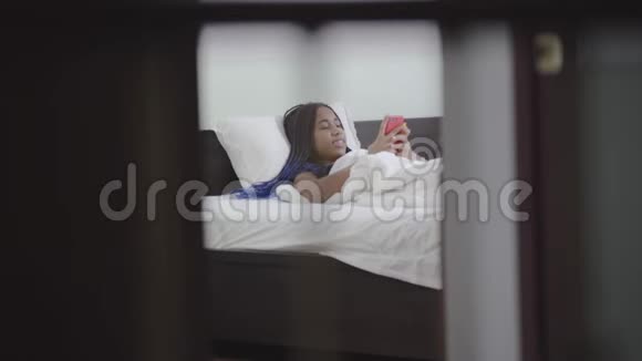 快乐微笑的非裔美国女孩与可怕的人使用智能手机在床上青少年晚上在她的网上冲浪视频的预览图