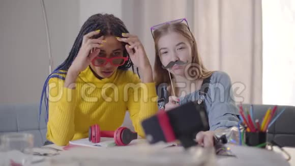 一位戴着红色眼镜的非裔美国女孩和她的白种人朋友坐在桌子旁看着假胡子视频的预览图