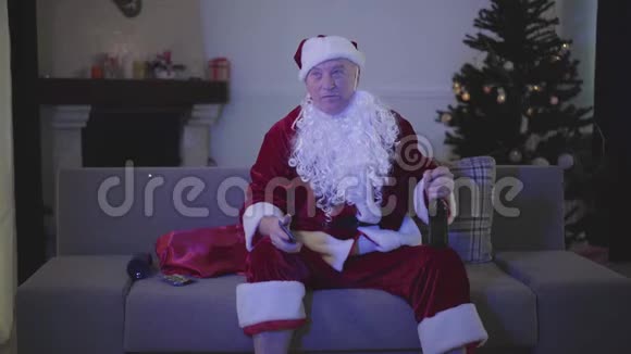 穿着圣诞老人服装的积极老人在家里坐在沙发上看电视喝啤酒高级官员视频的预览图