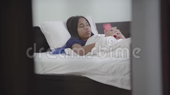体贴的女孩拿着智能手机打字和微笑的肖像非裔美国少年在白床上休息视频的预览图