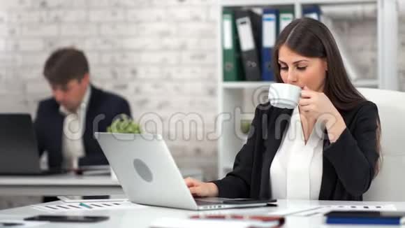 年轻女商人在工作中使用笔记本电脑时会在工作场所喝咖啡或茶视频的预览图