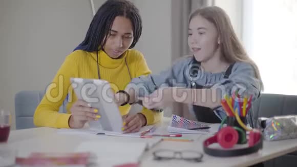 可爱的白种人女孩在平板电脑上展示非裔美国朋友的东西积极的学生在室内做作业视频的预览图